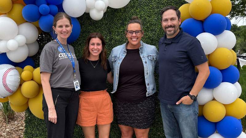 Four employees posing in front of a blue, yellow and white balloon arch